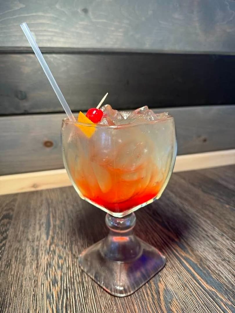 A vibrant red and orange cocktail with ice, a cherry, and an orange slice, on a wooden table.