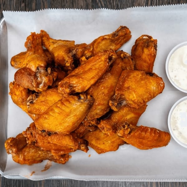 A tray of golden-brown chicken wings with two small cups of dipping sauce on white parchment paper.