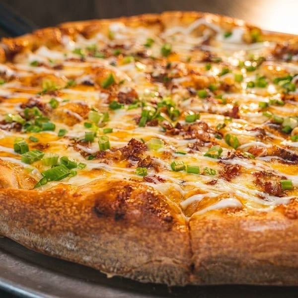 A whole pizza with melted cheese, bacon, green onions, and white sauce drizzle on a metal pan.