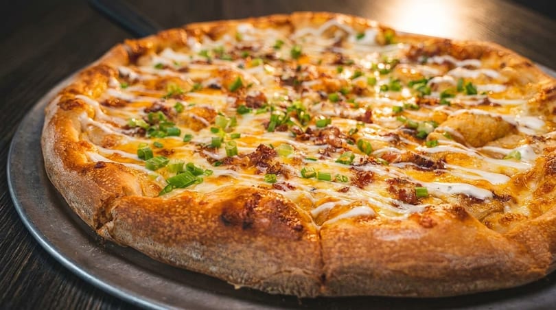 A whole pizza with melted cheese, bacon, green onions, and white sauce drizzle on a metal pan.