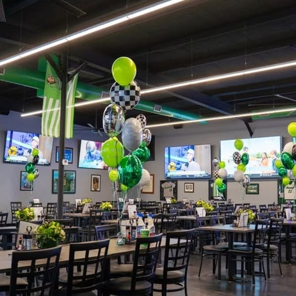 A restaurant dining room with tables, chairs, large TVs, and green, black, and white balloons.