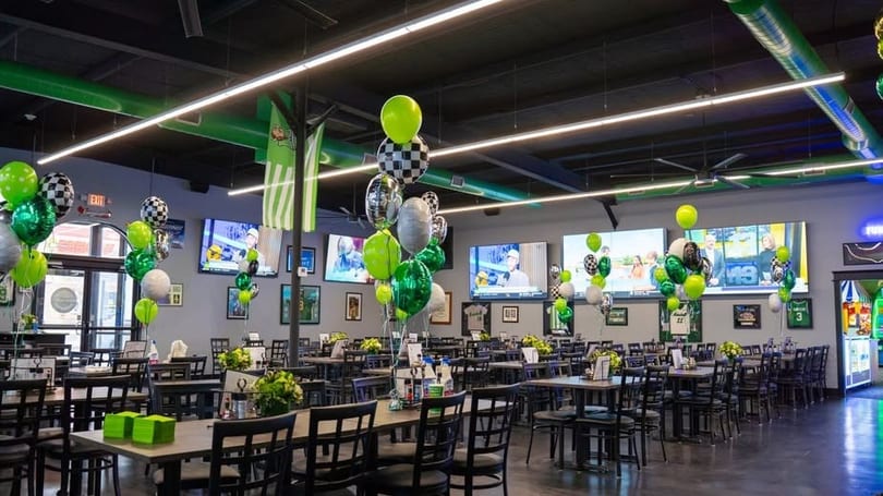 A restaurant dining room with tables, chairs, large TVs, and green, black, and white balloons.