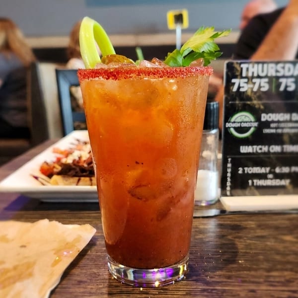 A Bloody Mary cocktail with celery and a red rim sits on a wooden table in a restaurant.