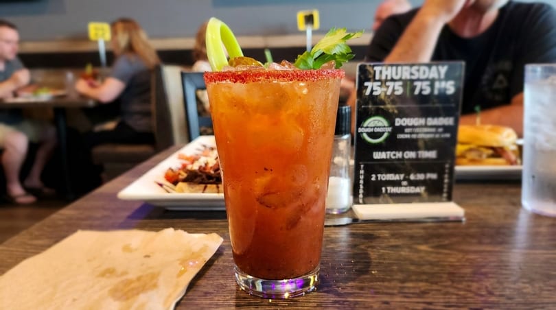 A Bloody Mary cocktail with celery and a red rim sits on a wooden table in a restaurant.