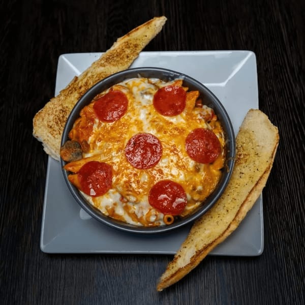 Baked pasta with melted cheese and pepperoni in a dark bowl, served with two slices of garlic bread.